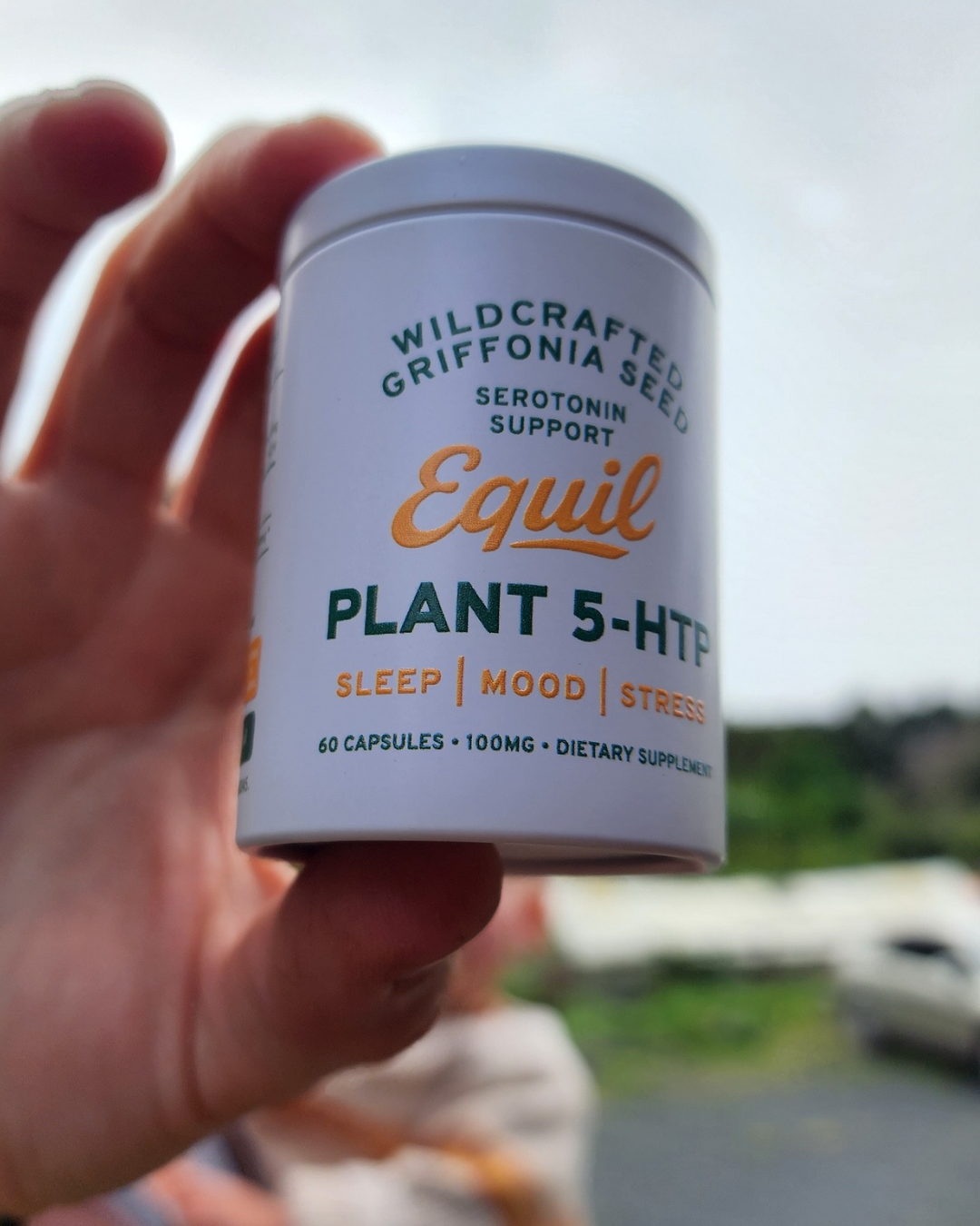 Hand holding a tub of Equil Plant 5-HTP capsules outdoors by the coast